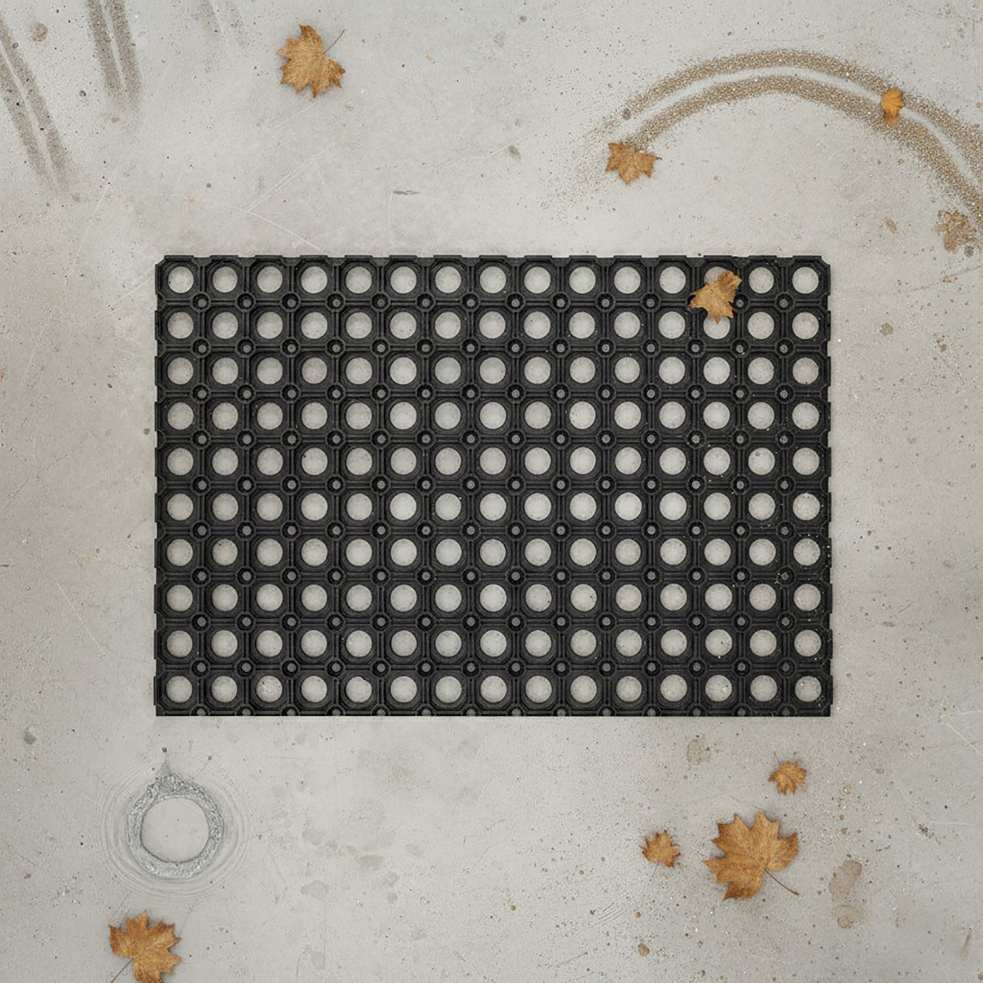 "Black rubber open-grid doormat on concrete surrounded by fallen autumn leaves and raindrops"