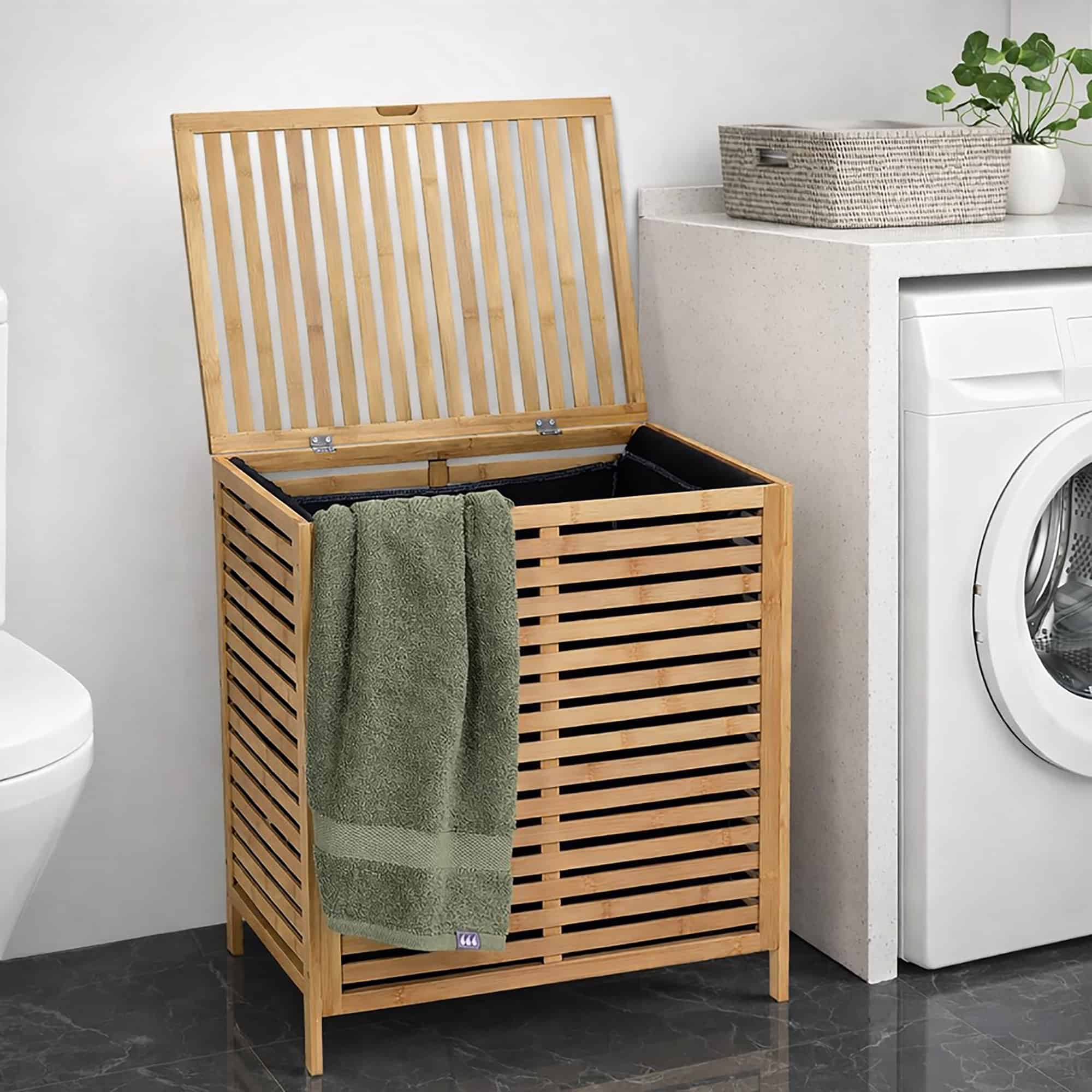 Bamboo slatted laundry hamper with lid open showing two-section interior with a green towel draped over the side, next to a washing machine
