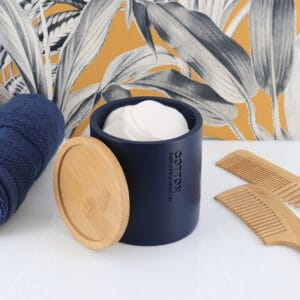 Open navy blue cotton jar with a bamboo lid, filled with cotton pads, styled with combs and rolled towels against a botanical wallpaper.