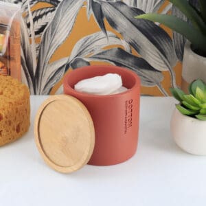 Open terracotta cotton jar with a bamboo lid, filled with cotton pads, styled with combs and rolled towels against a botanical wallpaper.