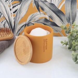 Open mustard yellow cotton jar with a bamboo lid, filled with cotton pads, styled with combs and rolled towels against a botanical wallpaper.