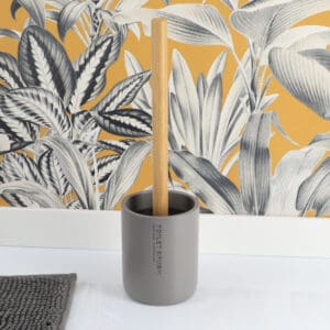 Gray toilet brush holder with a bamboo handle in front of a yellow leaf wallpaper.