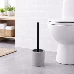 Minimalist gray toilet brush with black handle in a cylindrical concrete holder, placed on bathroom floor next to white toilet