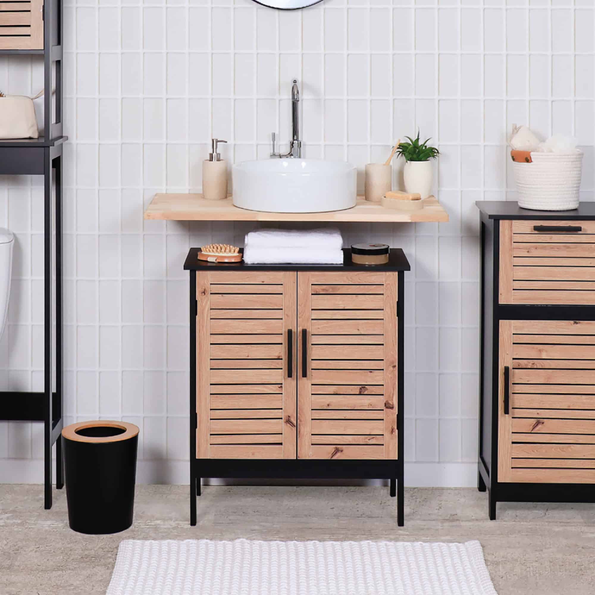 A stylish bathroom setting showcasing the cabinet under a round basin sink. The space is decorated with a wood shelf, toiletries, and a white tiled wall.