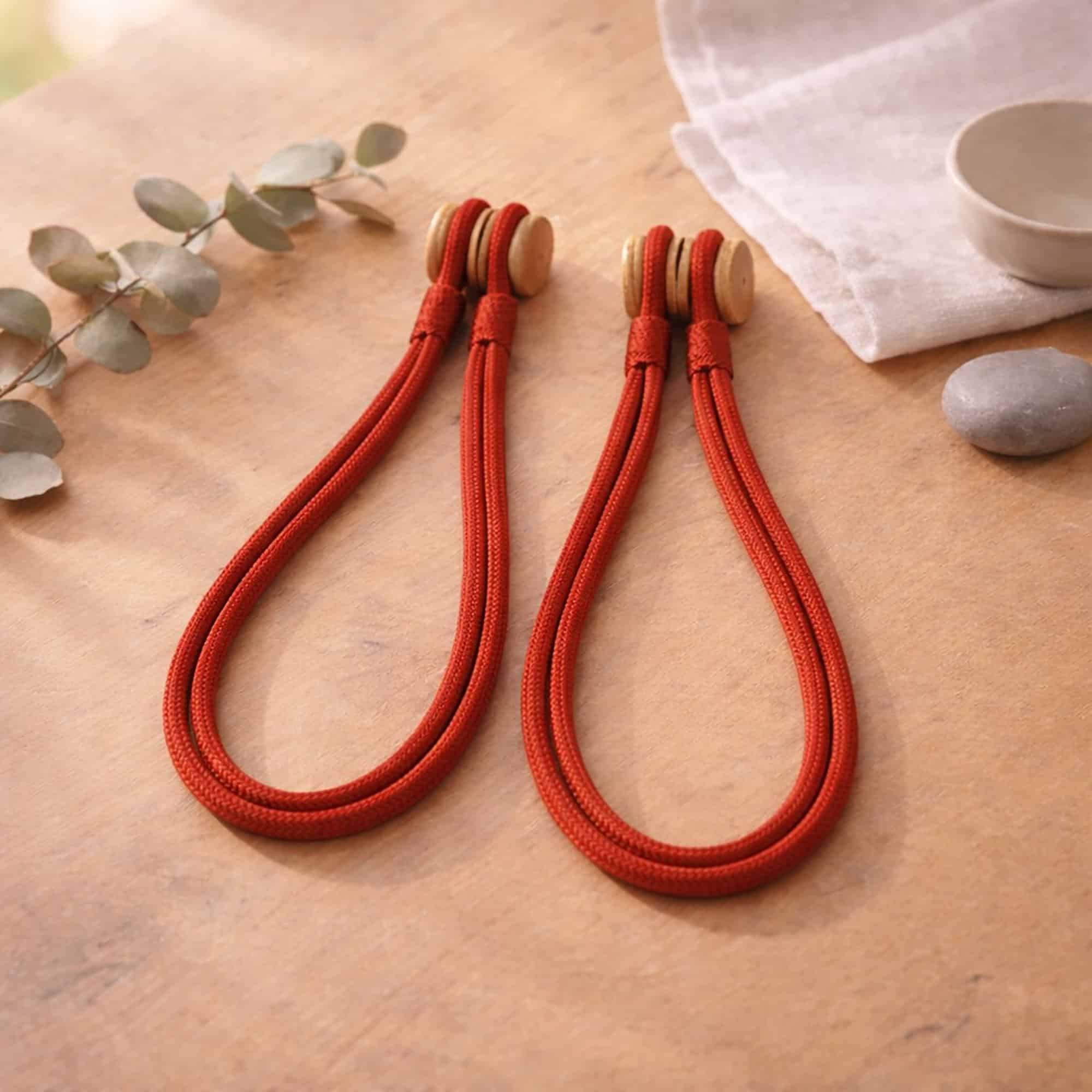 Rust red braided rope curtain tiebacks with a wooden magnetic closure, displayed on a wooden surface.