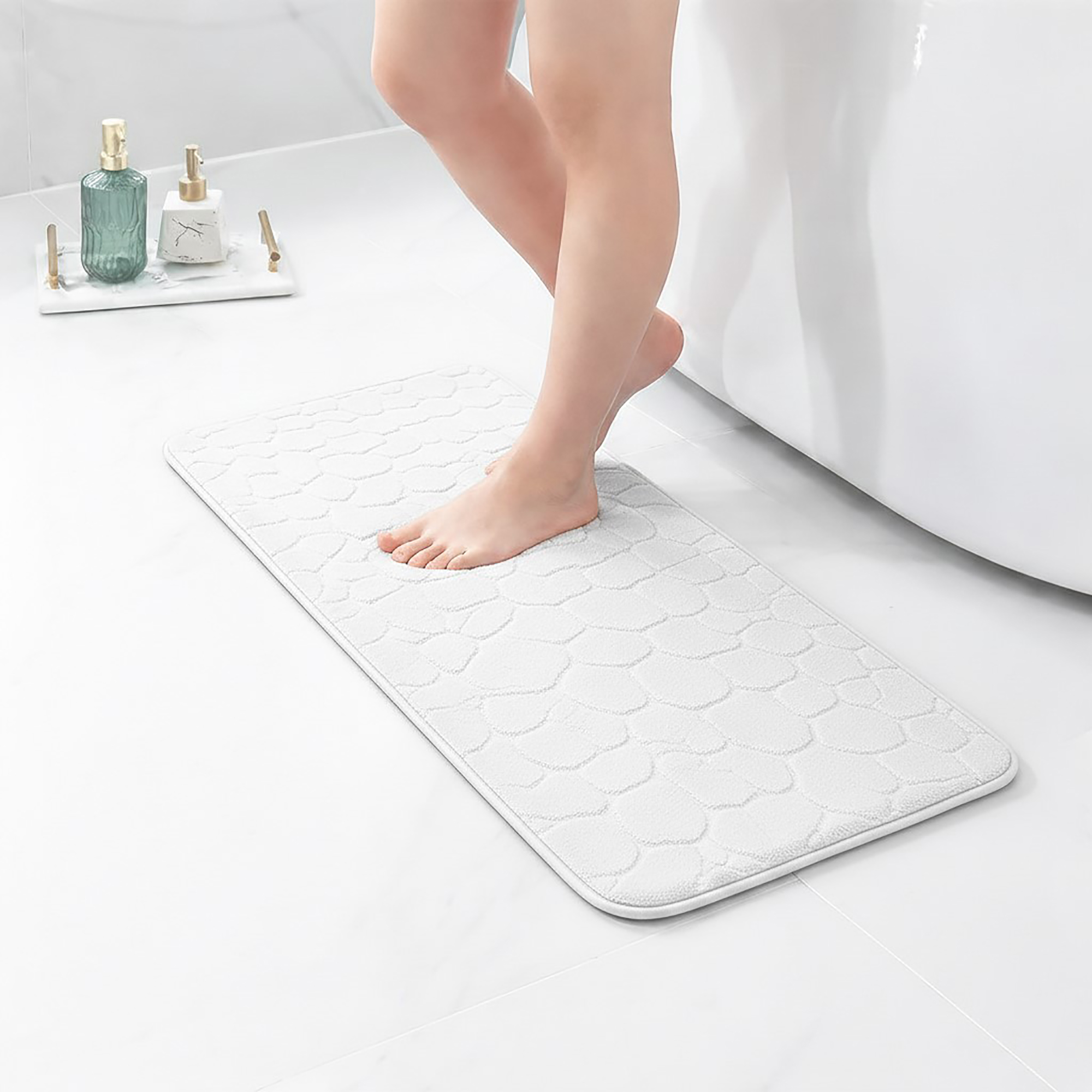 Barefoot stepping on a long white runner mat with pebble texture placed on a bathroom tile floor next to a tub
