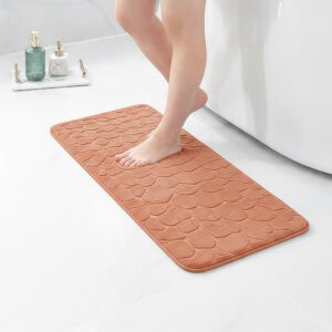 Barefoot stepping on a long copper runner mat with pebble texture placed on a bathroom tile floor next to a tub