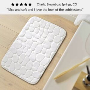 White bath mat with cobblestone pattern placed near sink on wood floor with customer review praising softness and design