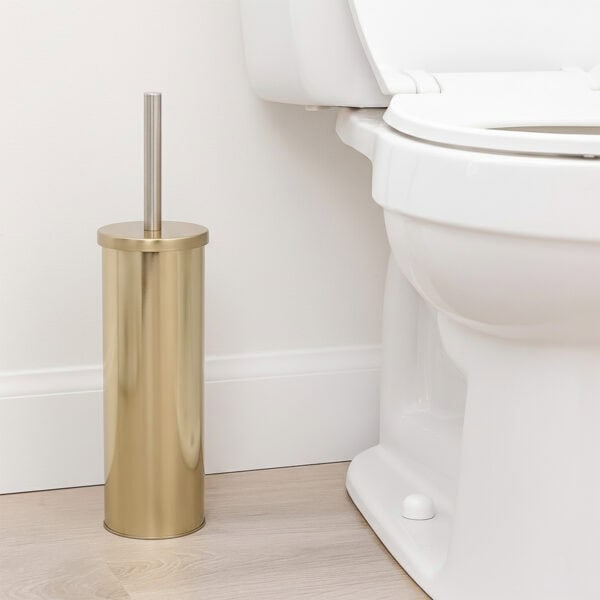 Brushed gold toilet brush with stainless steel handle in cylindrical holder beside white toilet