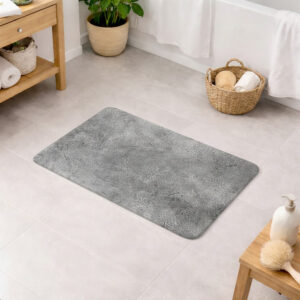 Gray bath mat with textured concrete pattern on a bathroom floor, surrounded by a wooden shelf, woven basket with bath accessories, bathtub, and soap dispenser with brush.