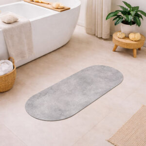 Oval runner bath mat with gray concrete texture on a bathroom floor next to a freestanding tub, woven stool with plant, wicker basket with towels, and jute rug.