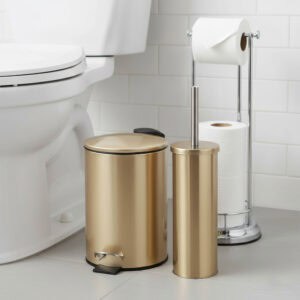 Brushed gold bathroom accessories set including step trash can, toilet brush holder, and chrome toilet paper holder stand in white tiled bathroom