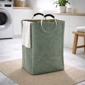 Sage green corduroy laundry basket with silver metal handles and a white towel draped over the side, in a bright laundry room.