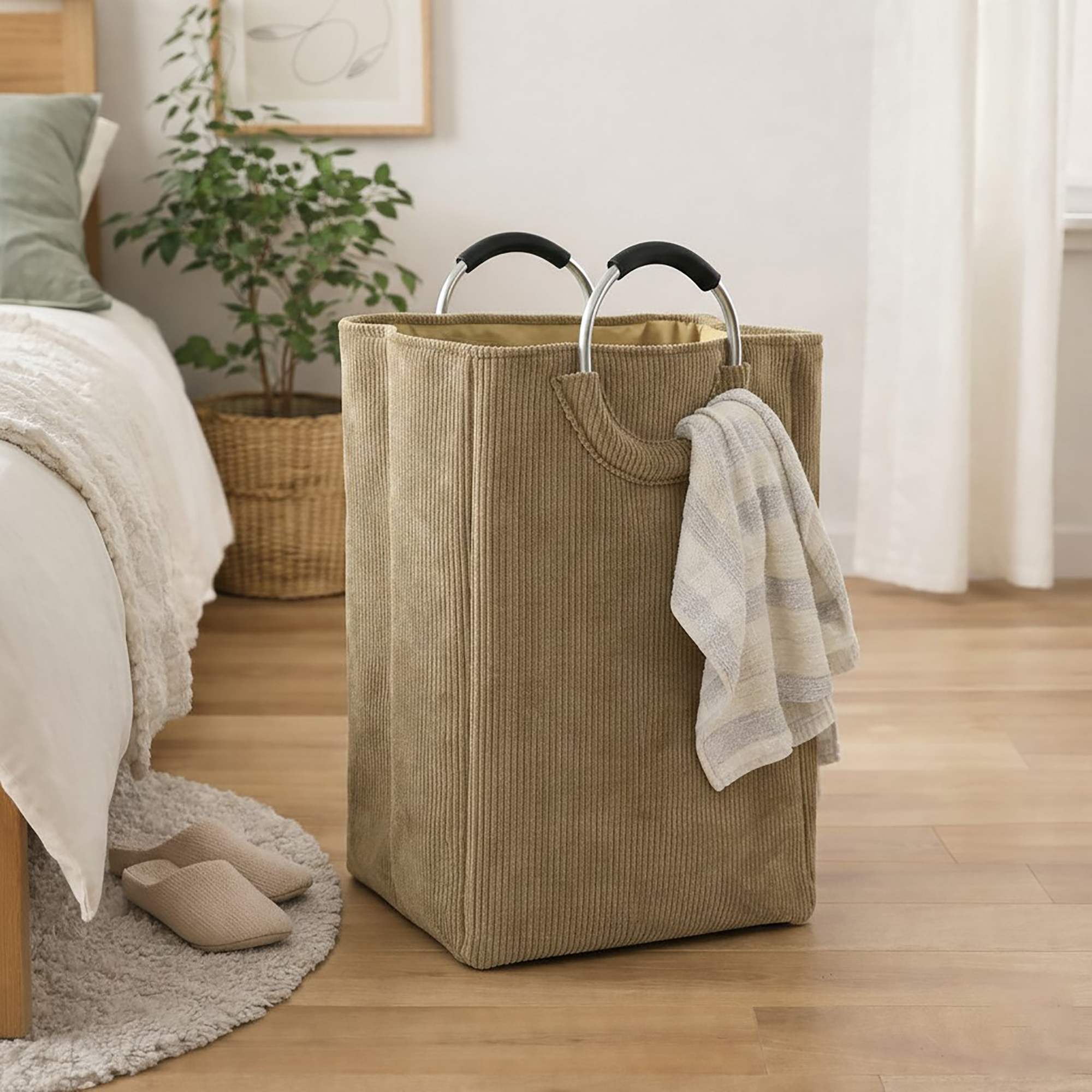 Sand beige corduroy laundry hamper with metal handles and a draped towel, styled in a cozy bedroom beside a bed and houseplant.