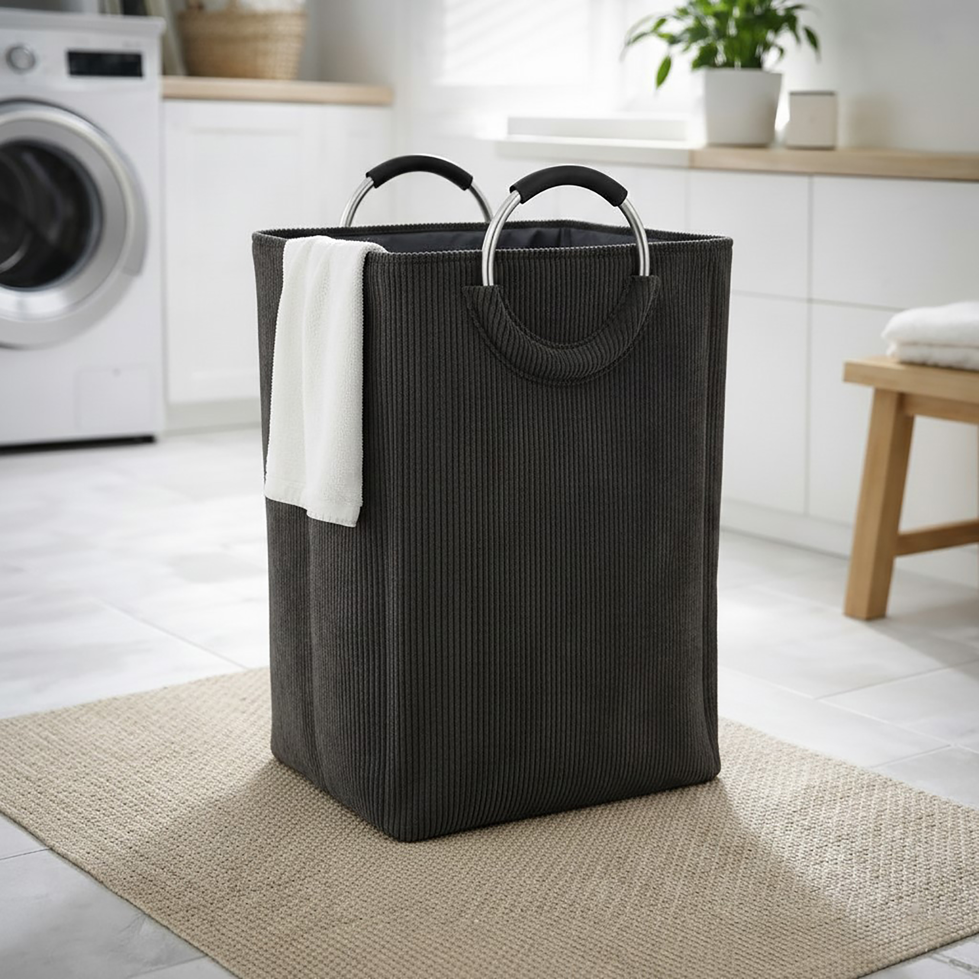 Charcoal gray corduroy laundry basket with silver metal handles and a white towel draped over the side, in a bright laundry room.