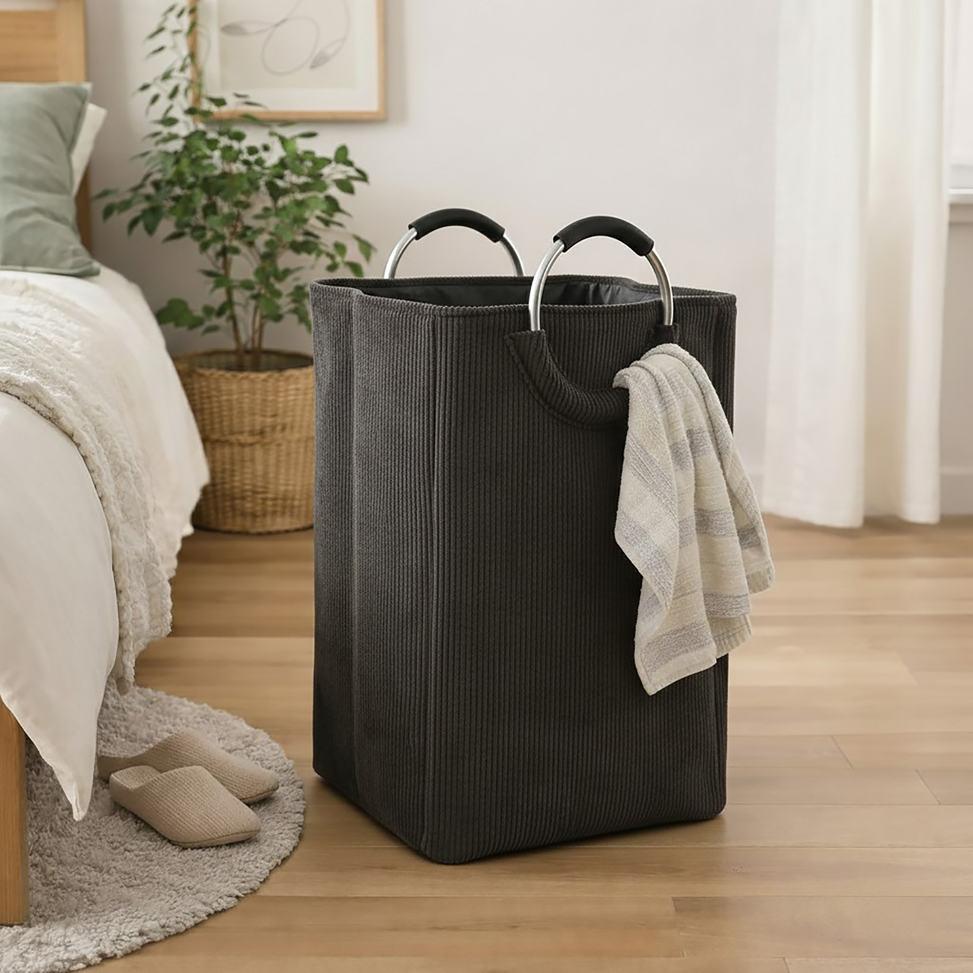 Charcoal gray corduroy laundry hamper with metal handles and a draped towel, styled in a cozy bedroom beside a bed and houseplant.