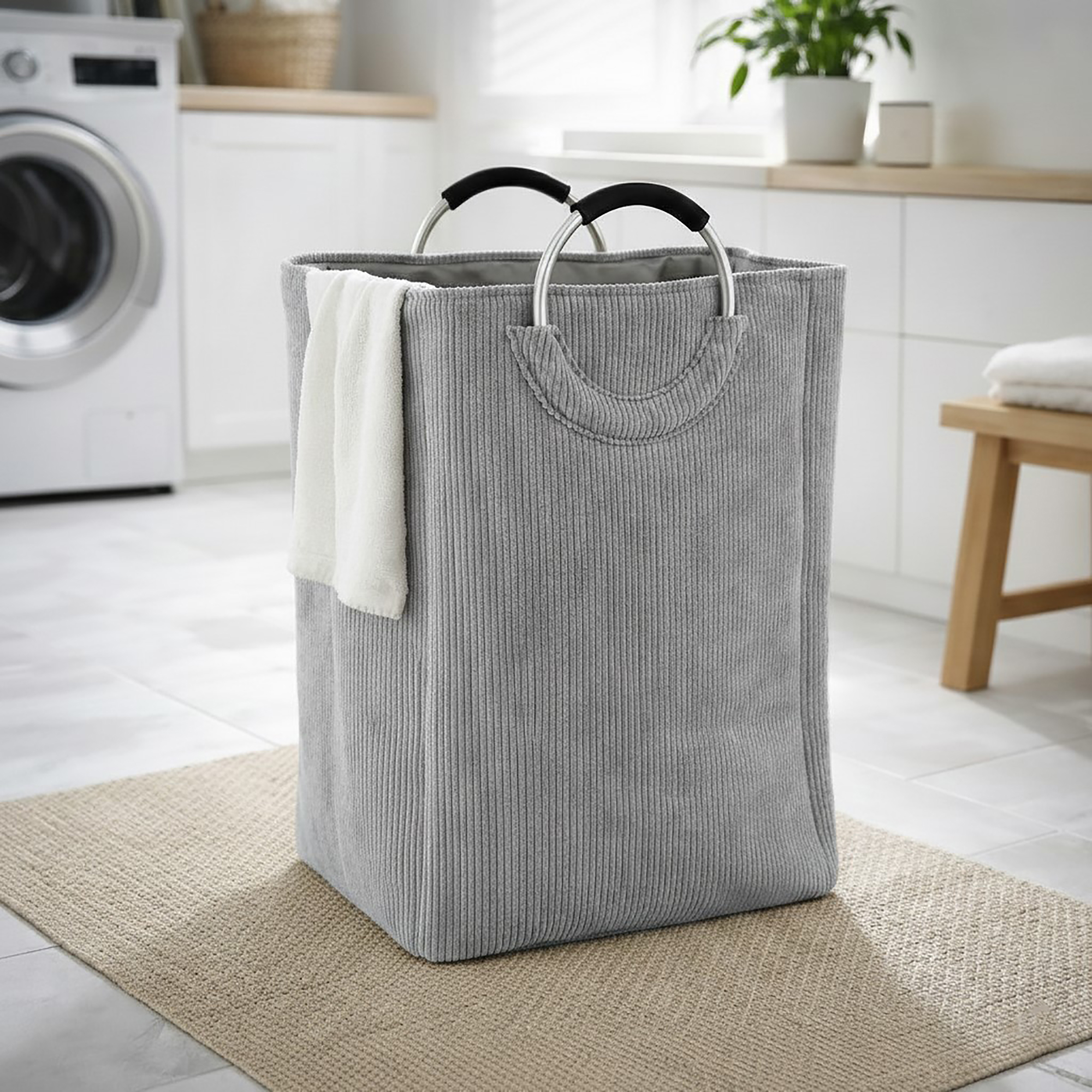 Soft gray corduroy laundry basket with silver metal handles and a white towel draped over the side, in a bright laundry room.