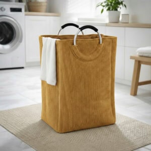 Mustard yellow corduroy laundry basket with silver metal handles and a white towel draped over the side, in a bright laundry room.