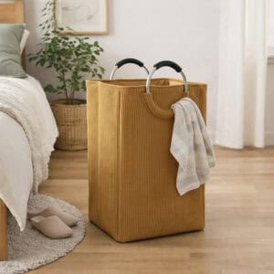 Mustard yellow corduroy laundry hamper with metal handles and a draped towel, styled in a cozy bedroom beside a bed and houseplant.