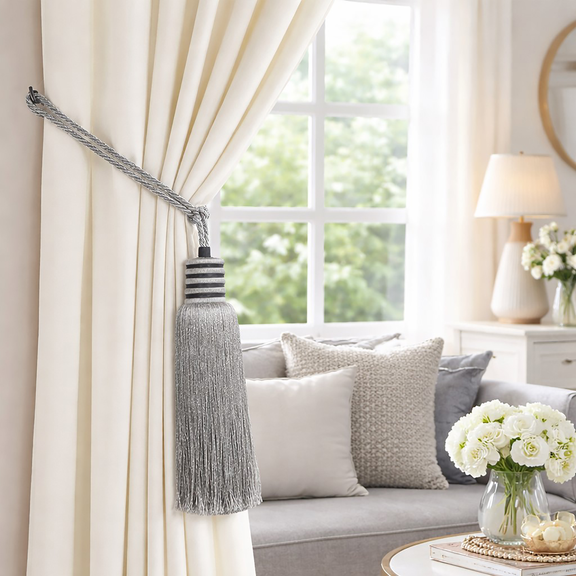 Silver mist tassel curtain tieback securing a cream drape beside a bright window with gray sofa in background
