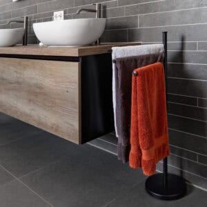 "Black 3-arm towel rack holding orange, gray, and white towels next to modern wood vanity"