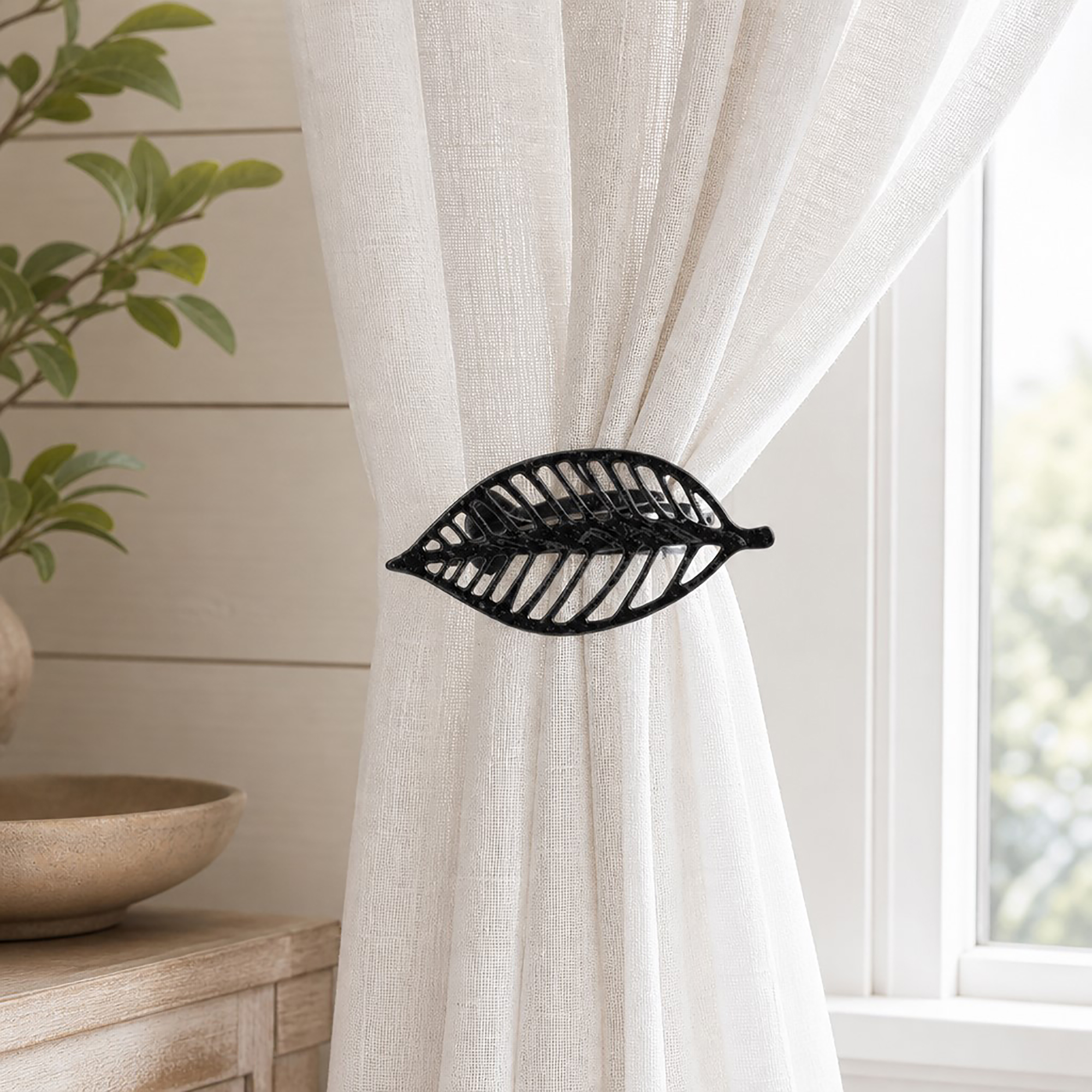 Textured black leaf curtain tieback holding back a white linen sheer panel by a window