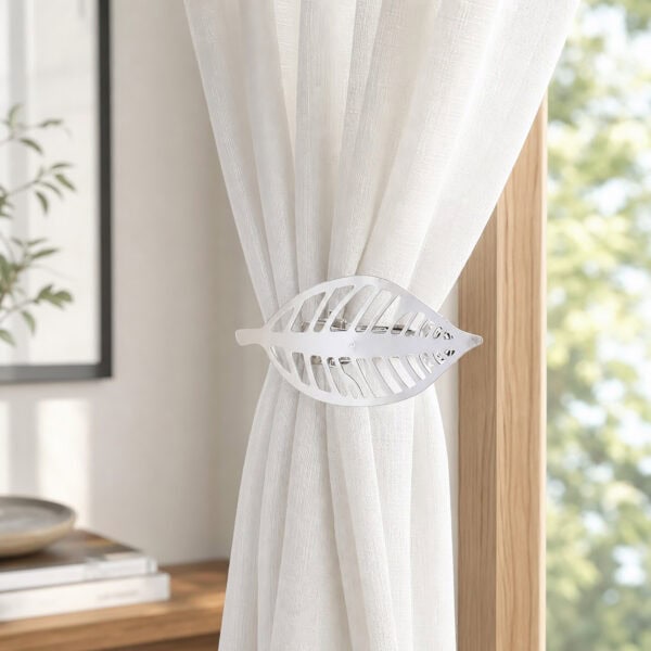 Polished silver leaf curtain tieback holding back a white sheer panel against a wood frame