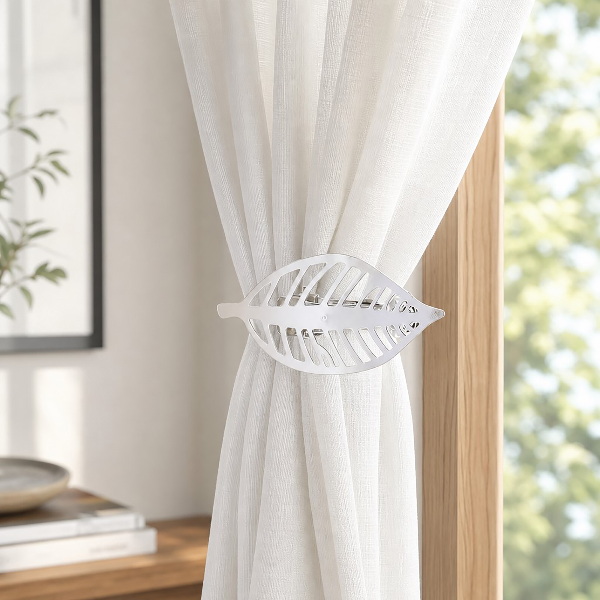 Polished silver leaf curtain tieback holding back a white sheer panel against a wood frame