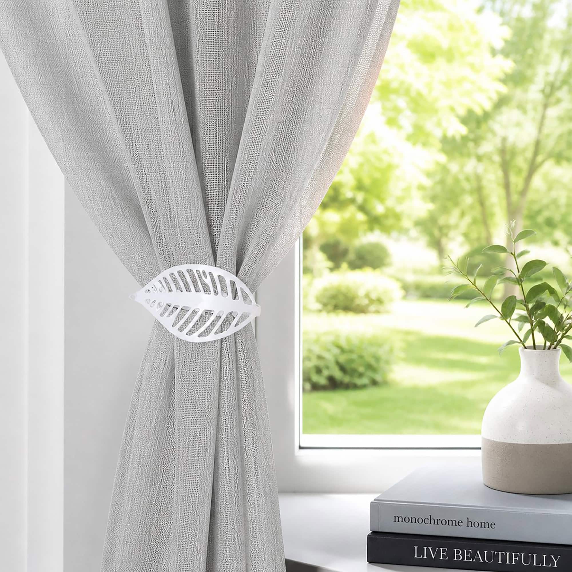 Polished silver leaf curtain tieback on a gray sheer panel by a sunny window with a vase