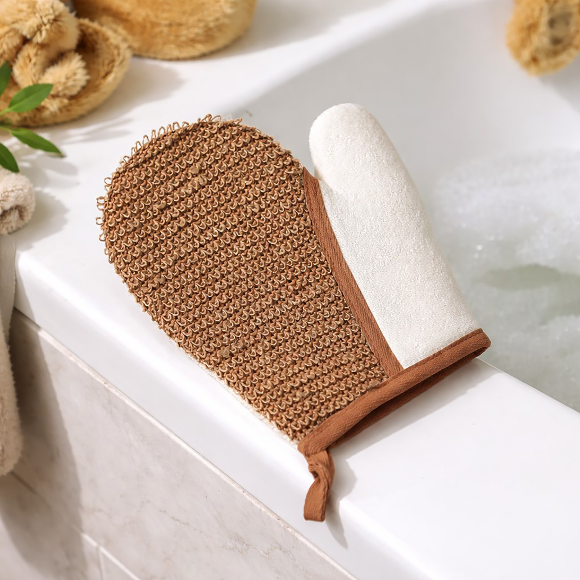 Dual-sided exfoliating mitt with textured brown side and white terry cloth side on bathroom tray