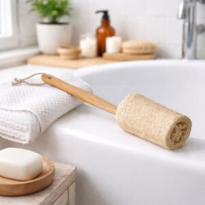 Natural loofah back scrubber with a wooden handle resting on a white bathtub ledge, with a folded towel, bar soap, and bath accessories nearby.