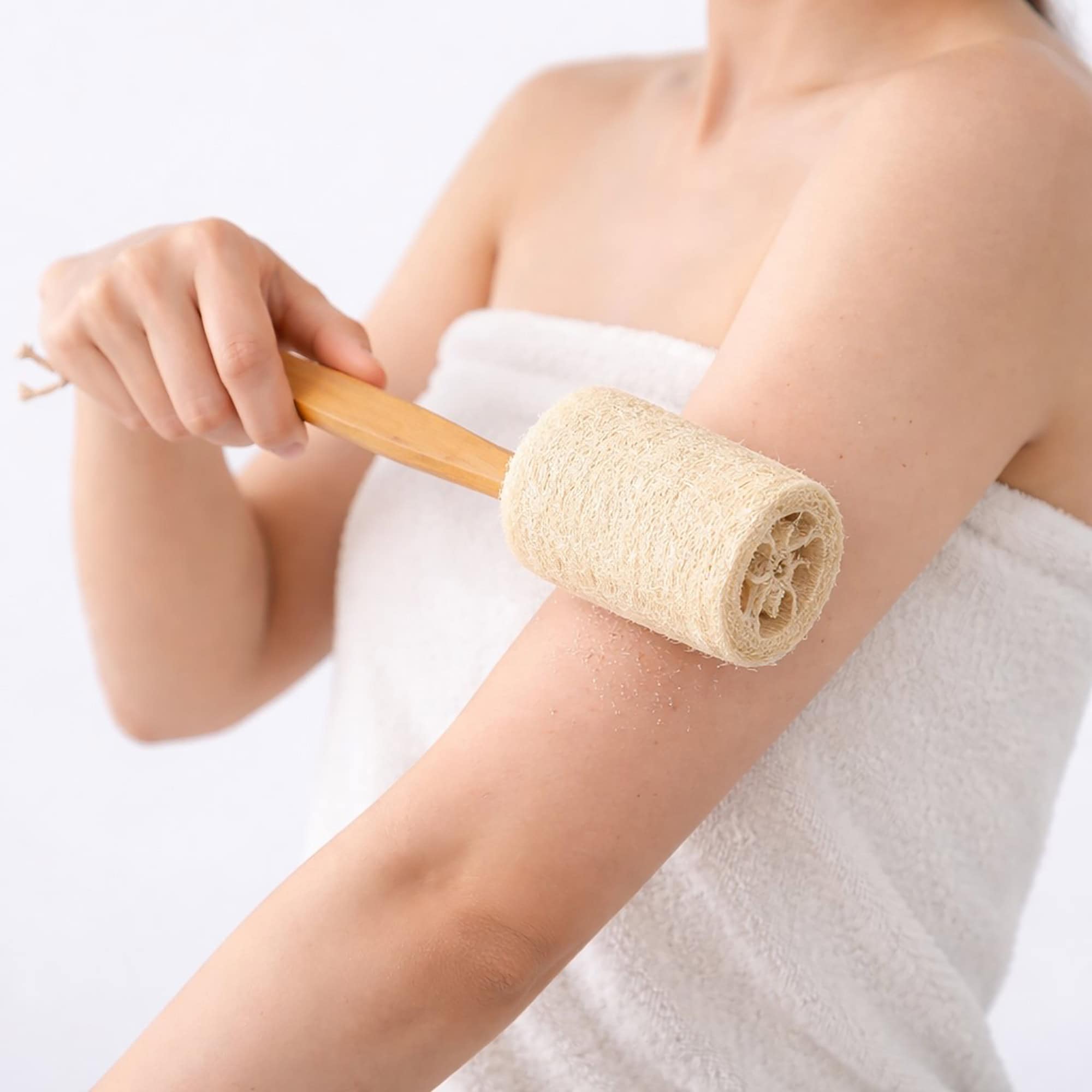 Woman in a towel using a long-handled natural loofah scrubber to exfoliate her arm.