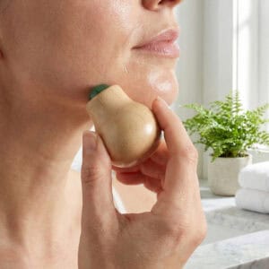 Hand using a wooden face massage roller with a jade stone ball along a woman's jawline in a bathroom setting.