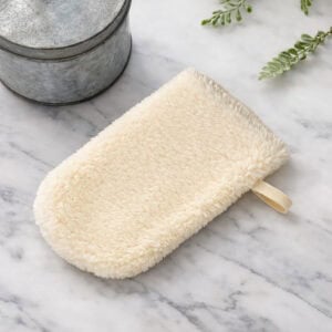 Cream-colored loofah mitt styled on white marble surface with metal containers and green plant sprigs