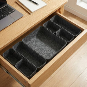 Seven-piece dark gray felt drawer organizer bins arranged empty in an open wooden desk drawer, with a laptop and notebook visible on the desktop.