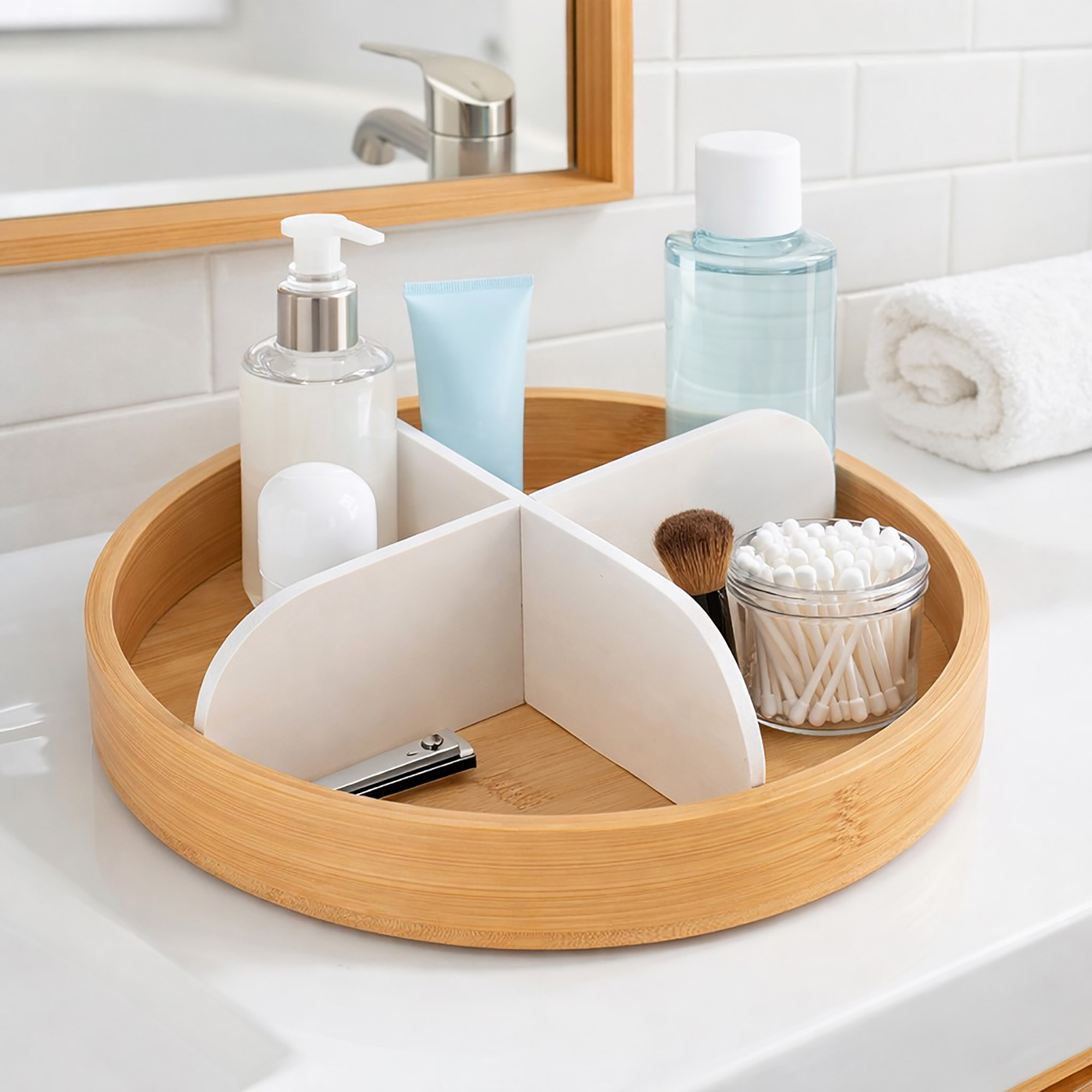 Round rotating bamboo tray with four white dividers, filled with skincare bottles, a makeup brush, nail clippers, and a glass jar of q-tips on a bathroom countertop.