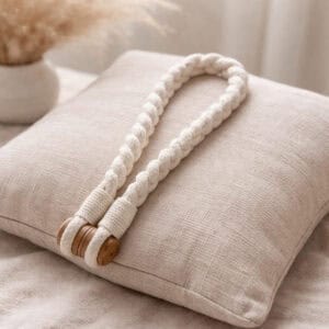 A twisted ivory cotton rope curtain tieback with a wooden button closure, draped over a cream linen cushion.