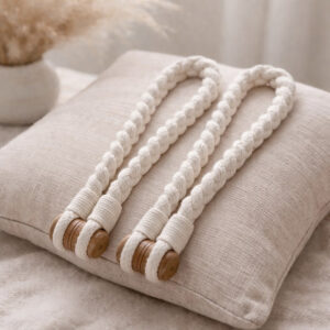 Twisted ivory cotton rope curtain tiebacks with a wooden button closure, draped over a cream linen cushion.