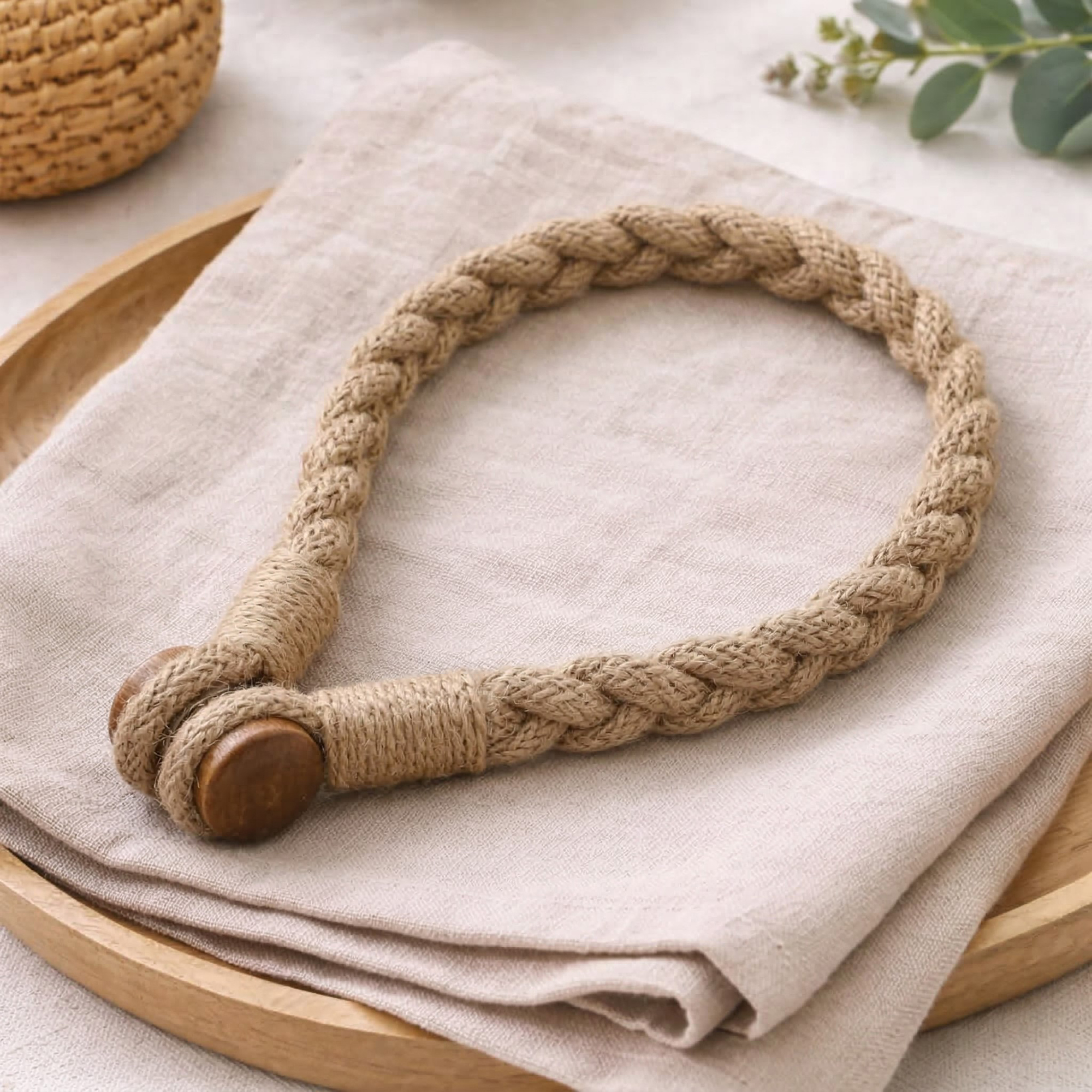A braided jute curtain tieback with a wooden button closure, laid on a linen cloth over a wooden tray.