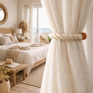 An ecru twisted rope curtain tieback with a wooden magnetic closure, holding back a sheer linen curtain in a coastal bedroom