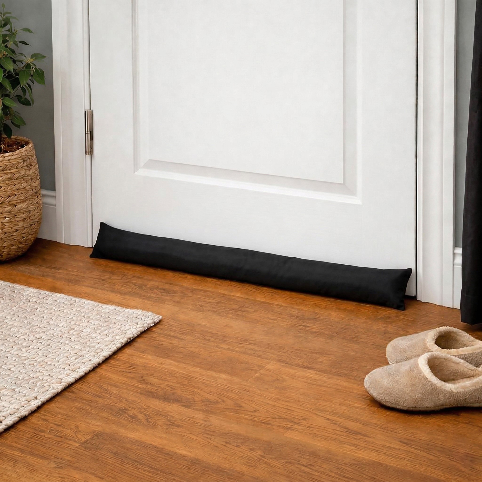 Draft stopper at a bedroom door base on wood flooring, next to a woven rug and a pair of slippers.