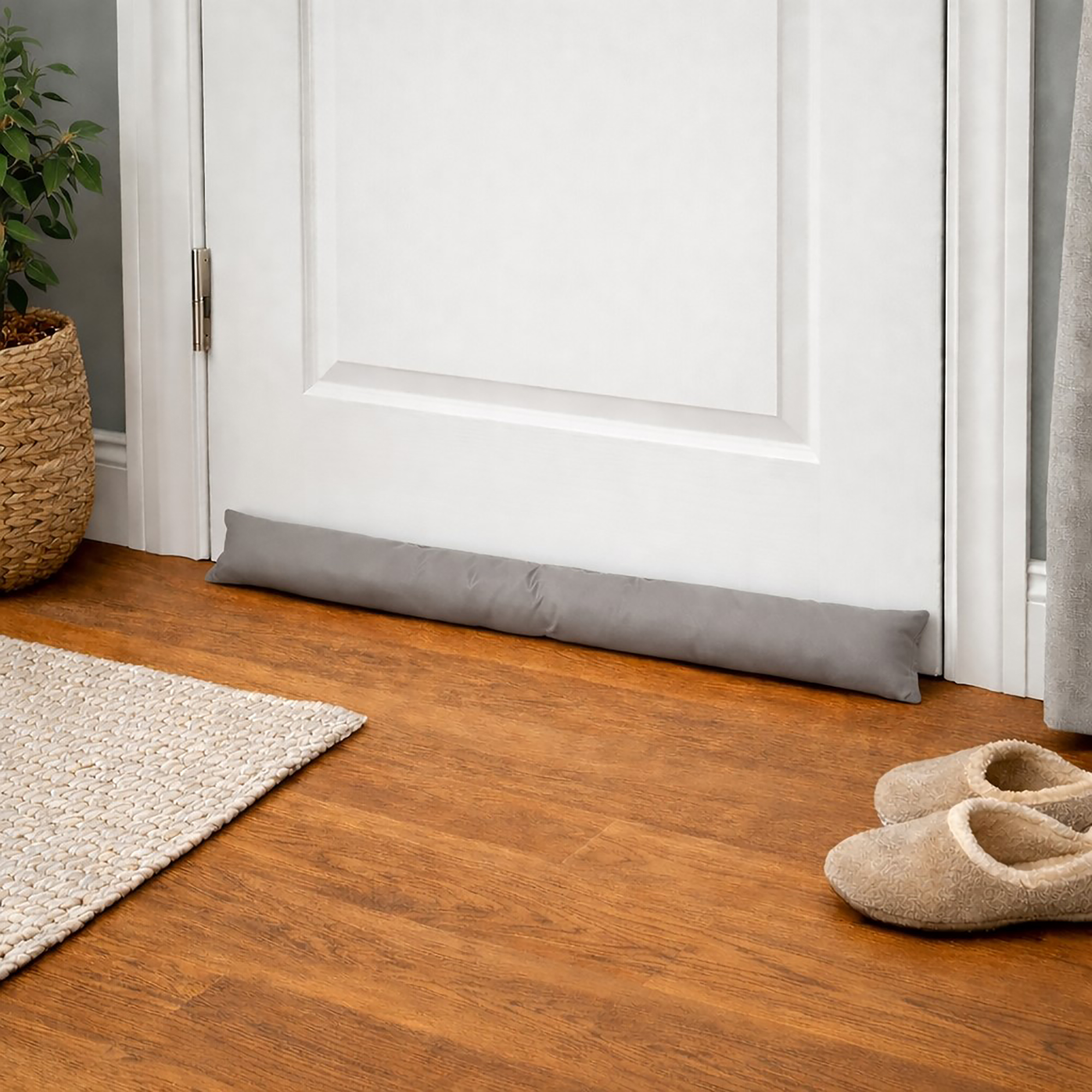 Draft stopper at a bedroom door base on wood flooring, next to a woven rug and a pair of slippers.