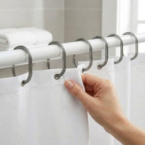Gray shower curtain hooks installed on white rod holding white curtain with hand adjusting fabric