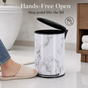 Bare foot in slipper presses the pedal to lift the lid on a white marble bathroom trash can, hands free operation beside a modern toilet.