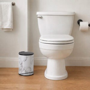 Small marble look bathroom trash can with black lid and pedal standing on wood floor next to a white toilet, slim profile for narrow spaces.