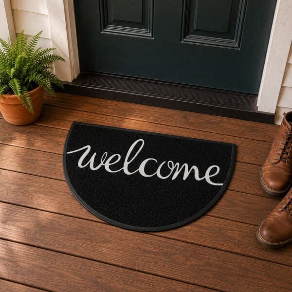 Black welcome mat with white cursive script displayed on a wood plank front porch beside a fern planter and brown leather boots, classic American home entrance