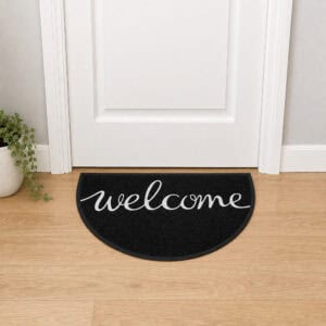 Black semi-circle welcome doormat with cursive script placed in front of a white interior door on light oak hardwood floor, modern minimalist entryway