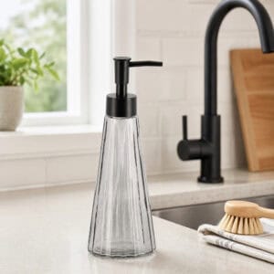 Smoked glass soap dispenser on a light kitchen counter near a matte black gooseneck faucet and natural sponge