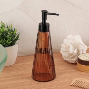 Amber glass soap dispenser styled with a green plant, white loofah, vanilla body scrub, and bamboo comb on a wood surface