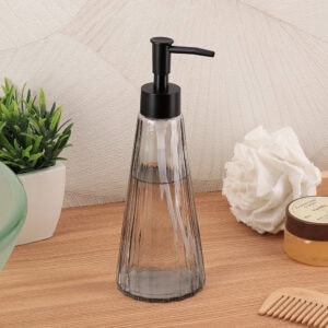 Smoked glass soap dispenser styled with a green plant, white loofah, vanilla body scrub, and bamboo comb on a wood surface
